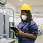 African American female factory worker in mask holding tablet
